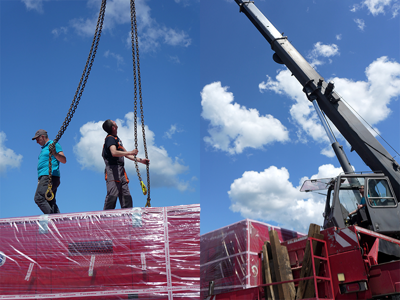 Two workers handling a red Zenessis generator wrapped in plastic, preparing for transport. One worker is guiding the equipment while the other stands nearby. The generator is suspended by a crane, with a clear sky and trees in the background.